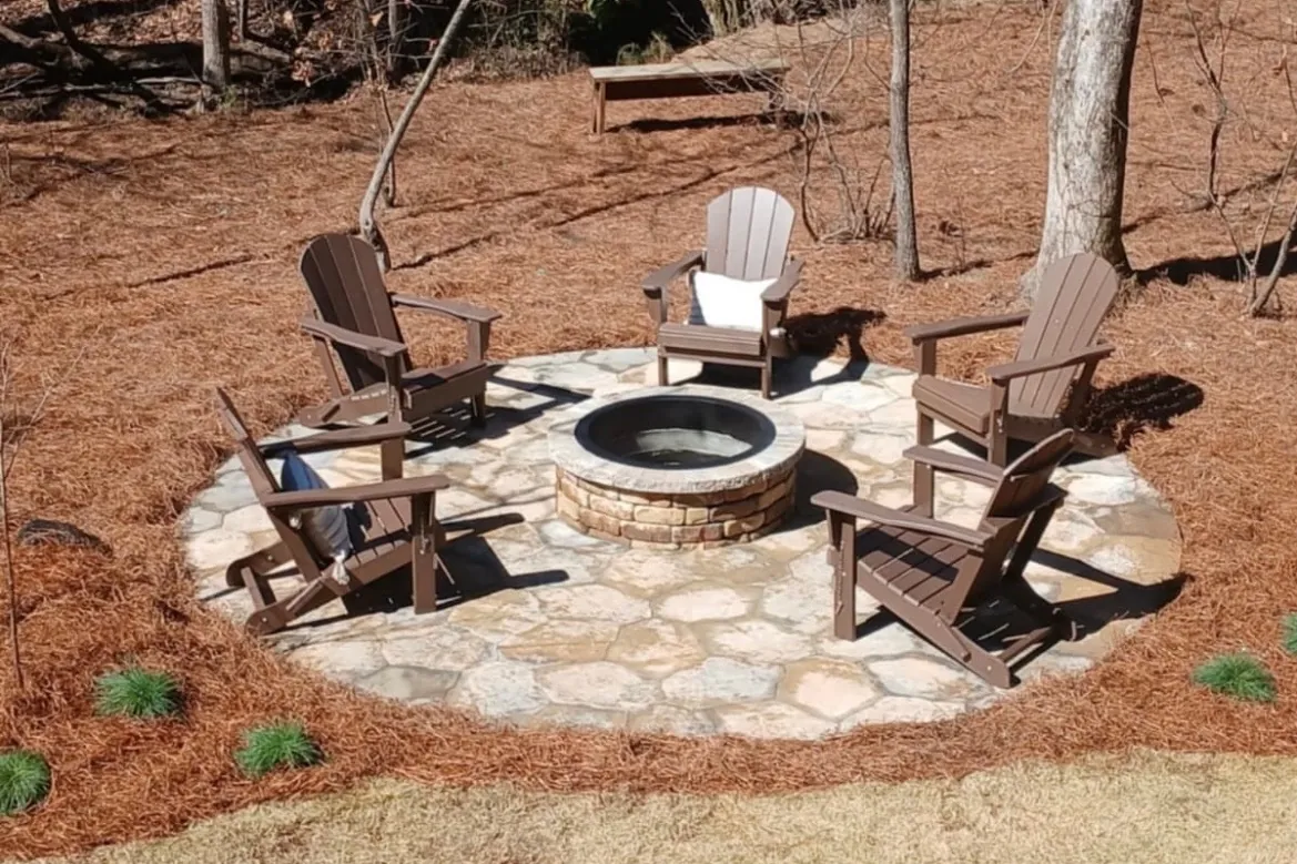 A round flagstone patio with a stone fire pit at its center, ringed by five Adirondack chairs in a wooded back yard covered in pine straw