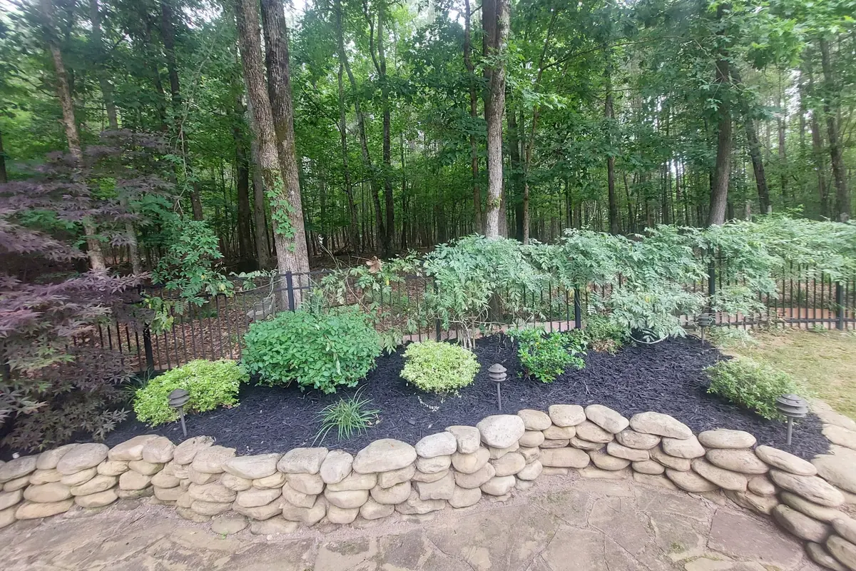A stacked stone border edging a black mulch bed of new plantings at a tree line