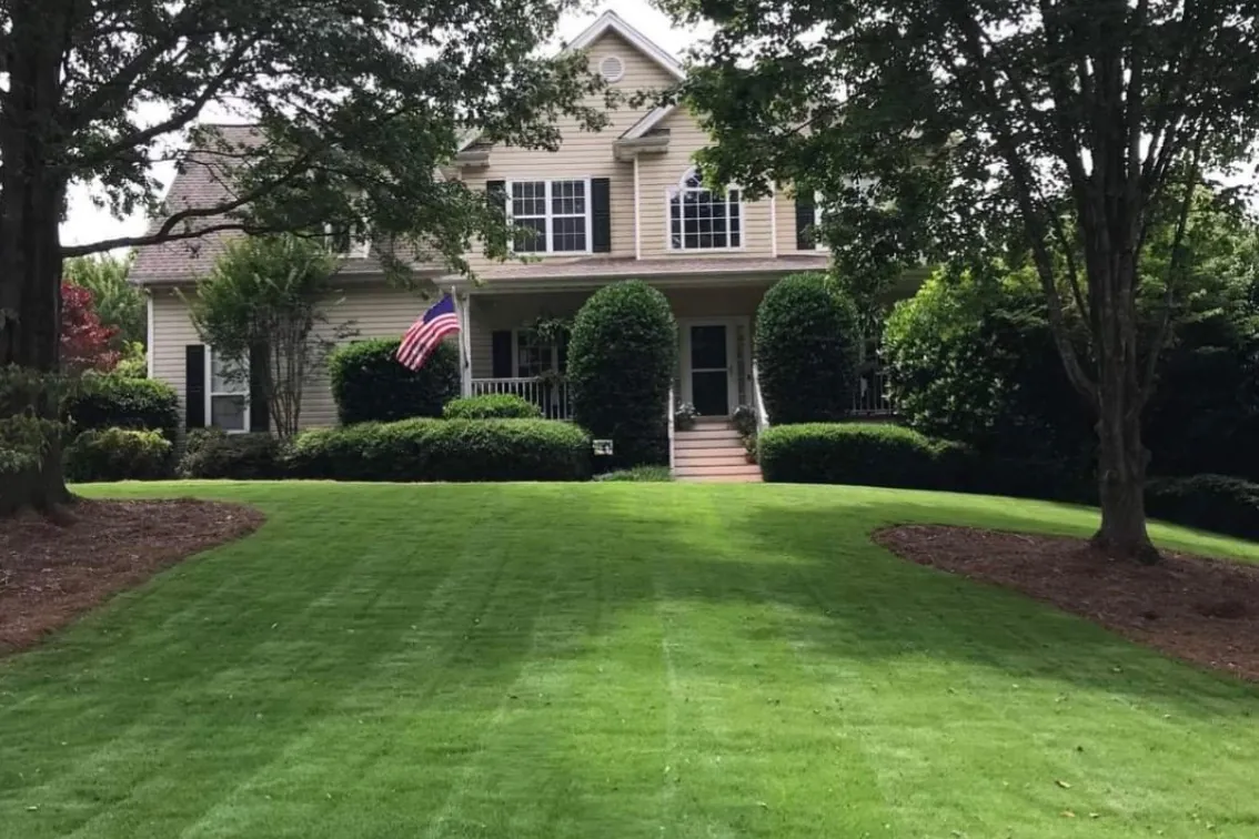 A two-story home with an American flag on the porch above a front lawn mowed in clean stripes, with mulched beds on both sides